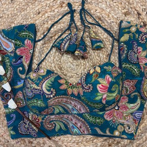 May include: Teal embroidered top with a floral and paisley pattern. The top is embellished with sequins and tassels. The fabric has a subtle sheen, and the design incorporates pink, purple, and green hues. The top is presented on a woven, natural-toned surface.
