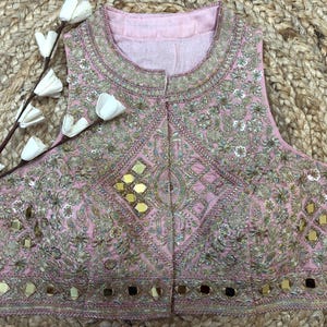 May include: A sleeveless, light pink top with intricate embroidery and mirror embellishments. The garment features a round neckline and a detailed pattern across the front. The design includes gold and silver threadwork, creating a luxurious look. The top is likely part of a formal wear collection.