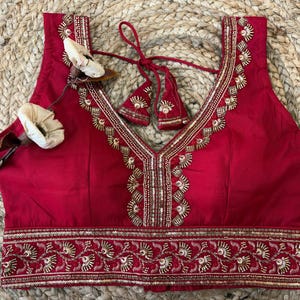 May include: A sleeveless, red blouse with intricate gold embroidery along the neckline and hem. The V-neck design is accentuated with delicate beadwork. The blouse features a tie closure and is adorned with floral embellishments.