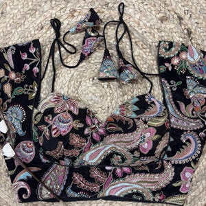 May include: Black velvet top with a colourful paisley pattern. The top has a sweetheart neckline, adjustable straps, and decorative tassels. The floral and paisley design includes pink, blue, and green accents.