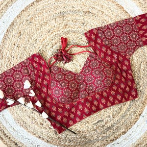 Puede incluir: Top rojo estampado con mangas largas y espalda anudada. La parte superior presenta un diseño floral repetido en un tono más claro de rojo y beige. La prenda se exhibe sobre una estera circular tejida con un borde blanco y una ramita de flores blancas.