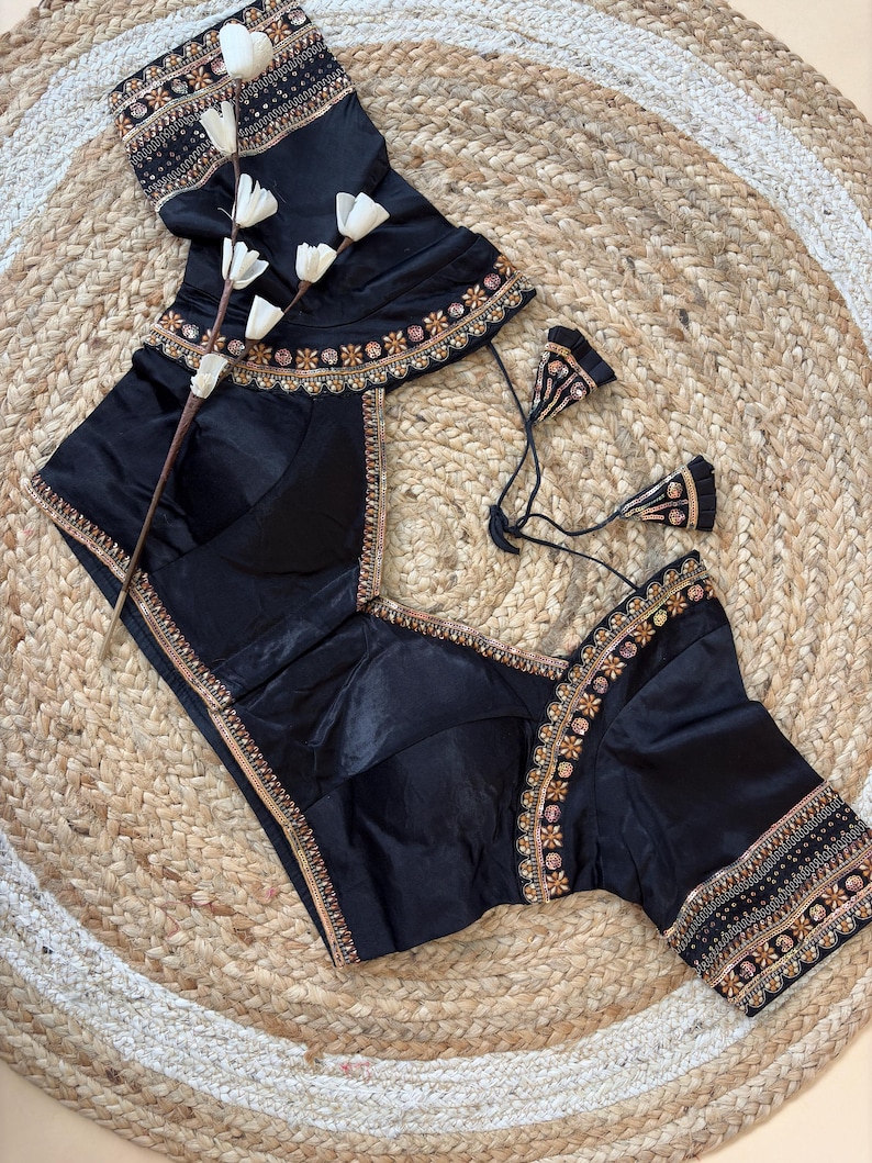 May include: Black silk blouse with intricate gold and pink embroidery along the neckline, sleeves, and hem. The blouse features a V-neck design and short sleeves. A matching black skirt is also visible.