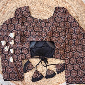 May include: A long-sleeved blouse with a black and brown floral pattern. The blouse has a square neckline and a black lining. Decorative tassels hang from the front. The blouse is displayed on a woven, circular mat.