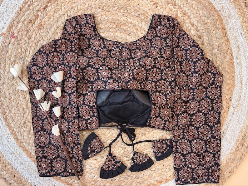May include: A long-sleeved blouse with a black and brown floral pattern. The blouse has a square neckline and a black lining. Decorative tassels hang from the front. The blouse is displayed on a woven, circular mat.