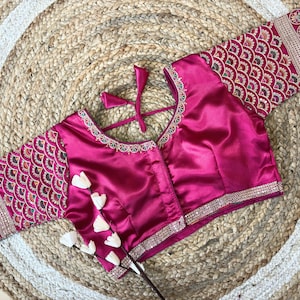 May include: A magenta-colored blouse with intricate embroidery on the sleeves and neckline. The sleeves feature a repeating pattern, and the neckline is trimmed with silver embellishments. The blouse has a tie closure at the back.