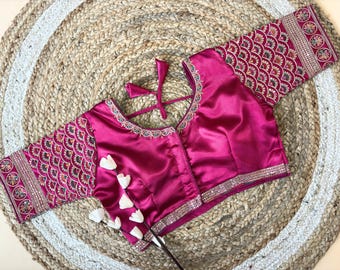 Può includere: Una camicetta color magenta con ricami intricati sulle maniche e sullo scollo. Le maniche presentano un motivo ripetuto e lo scollo è rifinito con abbellimenti argentati. La camicetta ha una chiusura con fiocco sul retro.