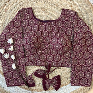 May include: A long-sleeved burgundy blouse with a repeating floral pattern in off-white. The blouse has a scoop neckline and a tie detail at the waist. The fabric appears to be a soft, lightweight material. The blouse is displayed on a woven, circular rug.
