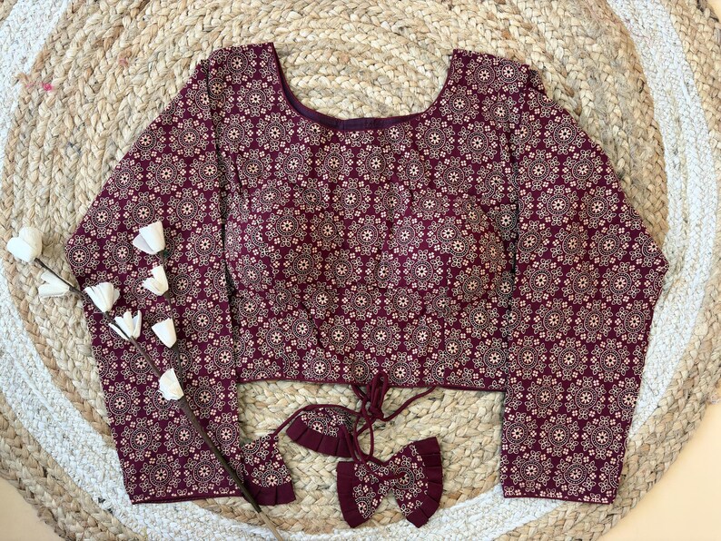May include: A long-sleeved burgundy blouse with a repeating floral pattern in off-white. The blouse has a scoop neckline and a tie detail at the waist. The fabric appears to be a soft, lightweight material. The blouse is displayed on a woven, circular rug.