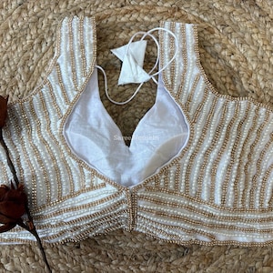 May include: Ivory-colored, sleeveless blouse with a V-neckline, adorned with rows of small, gold-colored beads. The blouse has a white lining and a tie closure at the back. The garment is displayed on a woven, beige surface.
