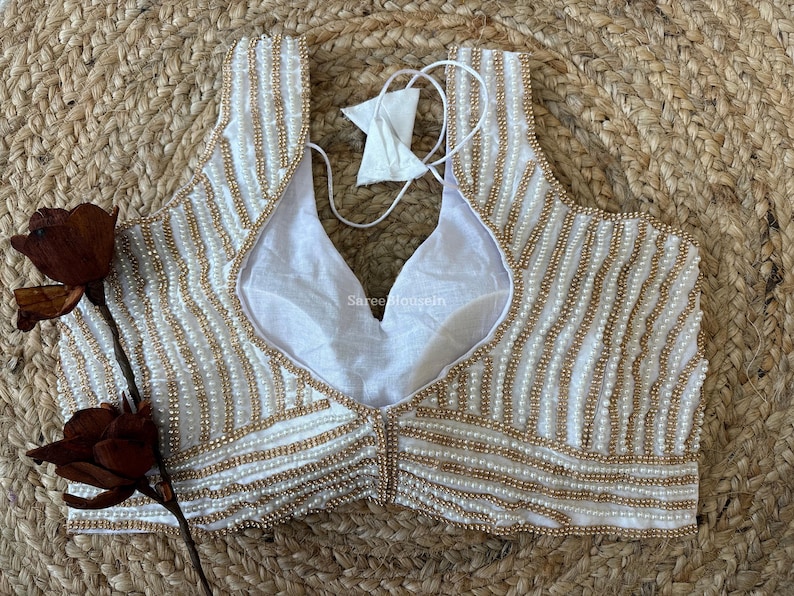 May include: Ivory-colored, sleeveless blouse with a V-neckline, adorned with rows of small, gold-colored beads. The blouse has a white lining and a tie closure at the back. The garment is displayed on a woven, beige surface.