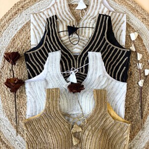 May include: Four sleeveless blouses with beaded embellishments in gold, black, white, and cream. The blouses have a V-neck design and are arranged on a woven mat. Each blouse has a tassel detail at the neckline.