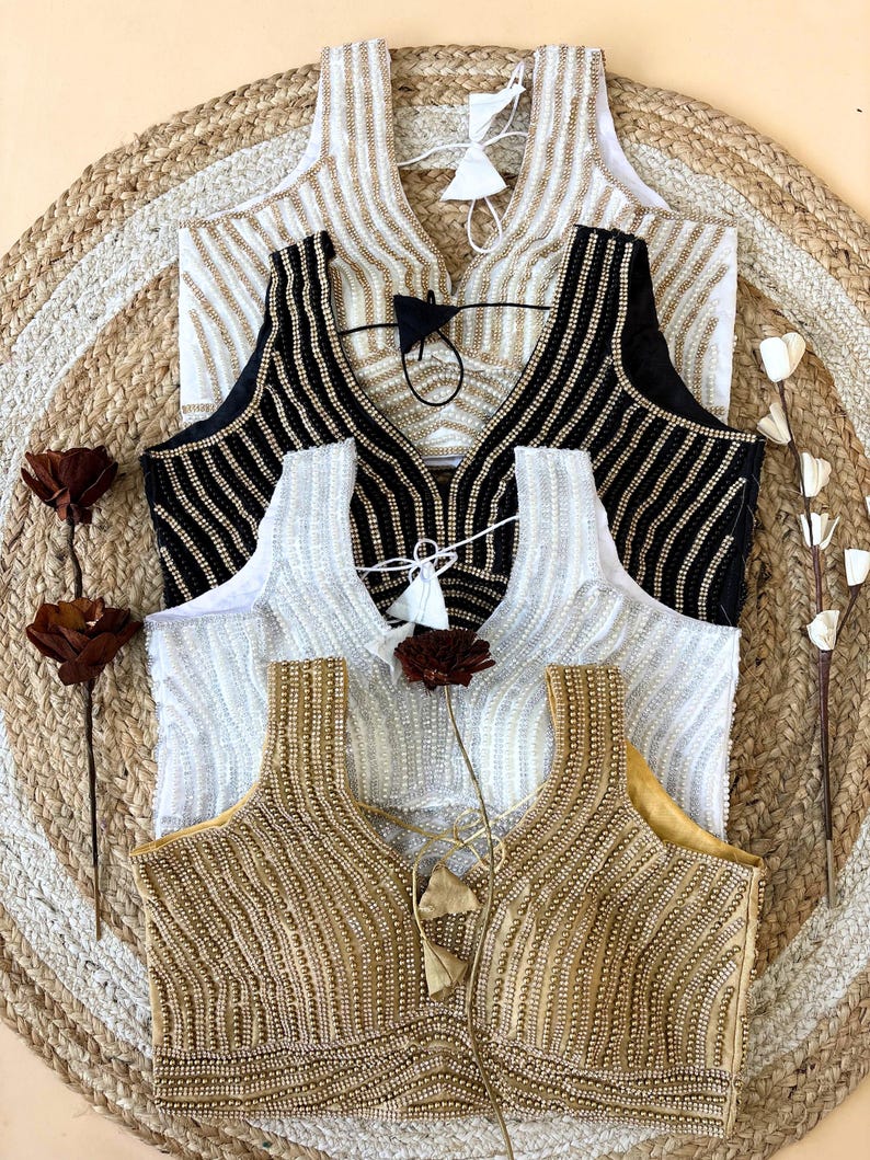 May include: Four sleeveless blouses with beaded embellishments in gold, black, white, and cream. The blouses have a V-neck design and are arranged on a woven mat. Each blouse has a tassel detail at the neckline.