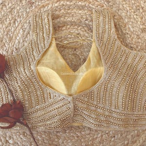 May include: A beige, beaded blouse with a deep V-neck and a light yellow lining. The blouse is adorned with rows of small, round beads. A dried, red rose is placed on the left side of the blouse. The background is a woven, tan surface.