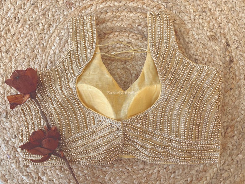 May include: A beige, beaded blouse with a deep V-neck and a light yellow lining. The blouse is adorned with rows of small, round beads. A dried, red rose is placed on the left side of the blouse. The background is a woven, tan surface.