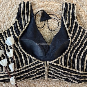 May include: Black sleeveless top with vertical rows of small, shiny beads. The top has a deep V-neck and a black lining. The design includes a decorative beaded pattern and a tie at the back. The background is a woven, natural fiber.