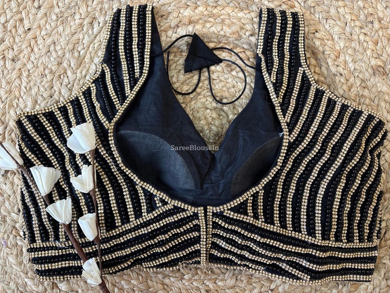 May include: Black sleeveless top with vertical rows of small, shiny beads. The top has a deep V-neck and a black lining. The design includes a decorative beaded pattern and a tie at the back. The background is a woven, natural fiber.