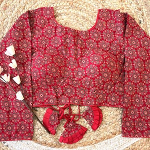 May include: A long-sleeved red blouse with a repeating circular pattern in a lighter shade of red and black. The blouse has a scoop neckline and decorative tassels at the waist. The garment is displayed on a woven, beige surface.