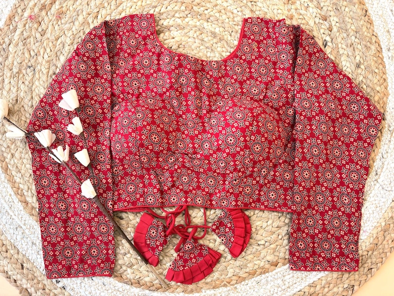 May include: A long-sleeved red blouse with a repeating circular pattern in a lighter shade of red and black. The blouse has a scoop neckline and decorative tassels at the waist. The garment is displayed on a woven, beige surface.