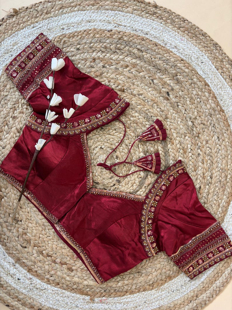 May include: A maroon blouse with intricate embroidery on the sleeves and neckline. The blouse features a square neckline and short sleeves. The design includes gold and cream-colored embellishments. The blouse is displayed on a woven, circular rug.