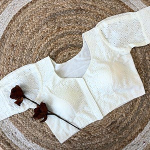 Może przedstawiać: Biała bluzka z ażurowym wzorem, krótkimi rękawami i dekoltem w serek. Bluzka wykonana jest z delikatnej, wzorzystej tkaniny. Na bluzce umieszczono suszoną, ciemnoczerwoną różę. Bluzka leży na okrągłym, tkanym dywanie jutowym.