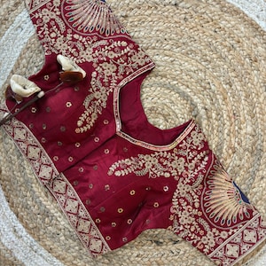 May include: A maroon blouse with intricate gold embroidery. The blouse features floral and geometric patterns, with a peacock feather design on the sleeves. The neckline and sleeve edges are trimmed with gold. The blouse is displayed on a woven mat.