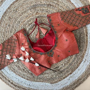 Può includere: Blusa color corallo con ricami intricati sulle maniche e sullo scollo. La blusa presenta uno scollo tondo con fodera rossa e nappe decorative. Le maniche sono decorate con motivi dettagliati e piccoli abbellimenti. La blusa è esposta su un tappeto intrecciato.