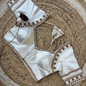 May include: Ivory-colored blouse with intricate embroidery along the neckline and sleeves. The blouse features a square neckline and short sleeves. The embroidery includes floral and geometric patterns in shades of brown and white.