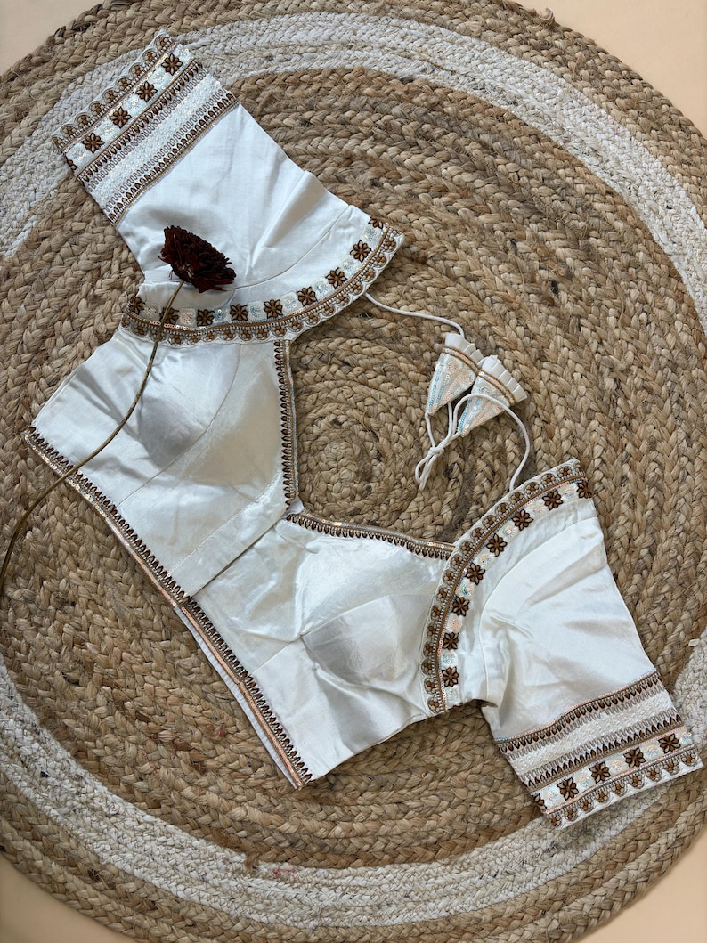 May include: Ivory-colored blouse with intricate embroidery along the neckline and sleeves. The blouse features a square neckline and short sleeves. The embroidery includes floral and geometric patterns in shades of brown and white.