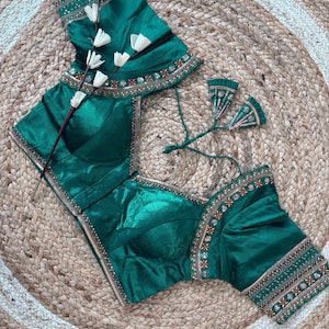 May include: Emerald green blouse with intricate embroidery along the neckline and sleeves. The blouse features a square neckline and short sleeves. The fabric has a subtle sheen. The blouse is displayed on a woven, circular rug.