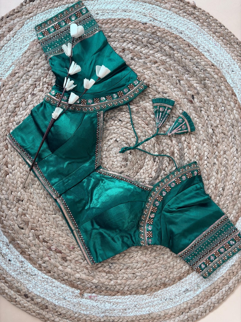 May include: Emerald green blouse with intricate embroidery along the neckline and sleeves. The blouse features a square neckline and short sleeves. The fabric has a subtle sheen. The blouse is displayed on a woven, circular rug.