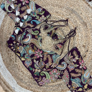 May include: A set of purple paisley-patterned clothing items, including a top and two matching pieces. The floral and paisley design features shades of pink, blue, and green. The items are displayed on a woven, circular surface.