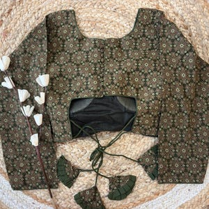 May include: A long-sleeved blouse with a dark green and gold floral pattern. The blouse has a scoop neckline and a tie closure in the back. The sleeves are wide and flared. The blouse is displayed on a woven surface.