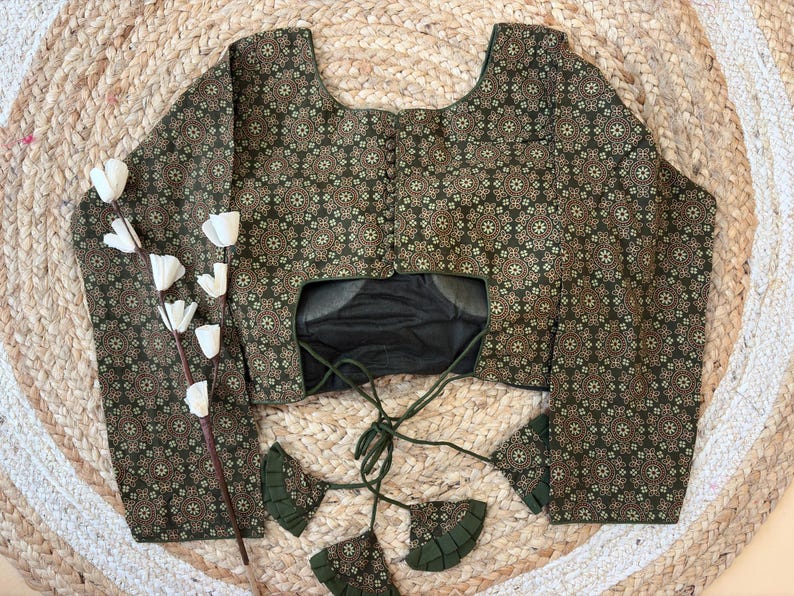 May include: A long-sleeved blouse with a dark green and gold floral pattern. The blouse has a scoop neckline and a tie closure in the back. The sleeves are wide and flared. The blouse is displayed on a woven surface.