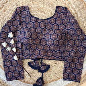 May include: A long-sleeved blouse with a navy blue background and a repeating pattern of small, stylized floral shapes in shades of brown and white. The blouse has a scoop neckline and decorative ties at the waist.