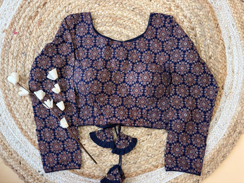 May include: A long-sleeved blouse with a navy blue background and a repeating pattern of small, stylized floral shapes in shades of brown and white. The blouse has a scoop neckline and decorative ties at the waist.
