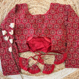 May include: A red blouse with a detailed floral pattern. The long-sleeved top features a scoop neckline and decorative tassels. The blouse is displayed on a woven surface, with white floral accents.