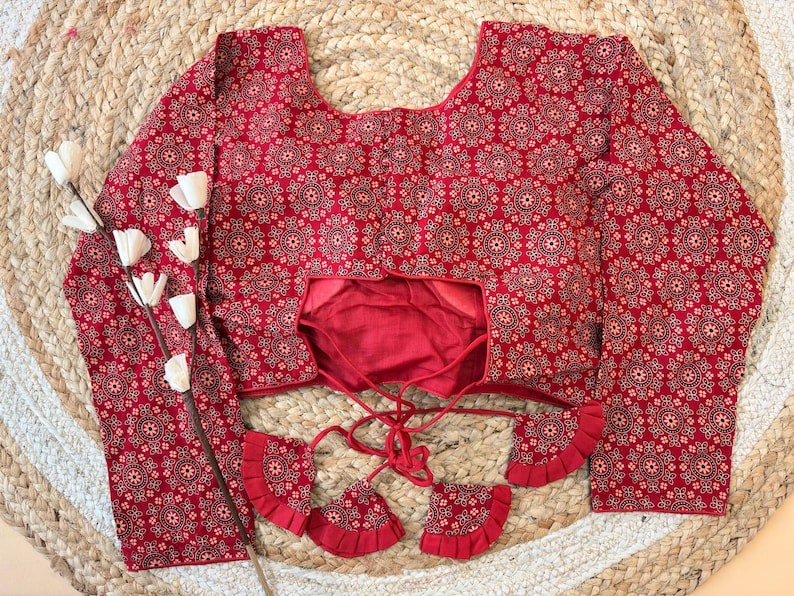 May include: A red blouse with a detailed floral pattern. The long-sleeved top features a scoop neckline and decorative tassels. The blouse is displayed on a woven surface, with white floral accents.