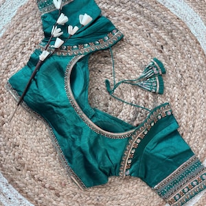 May include: Emerald green blouse with intricate embroidery along the neckline and sleeves. The blouse features a round neckline and short sleeves. The embroidery includes gold and copper-colored accents. The blouse is displayed on a woven jute rug.