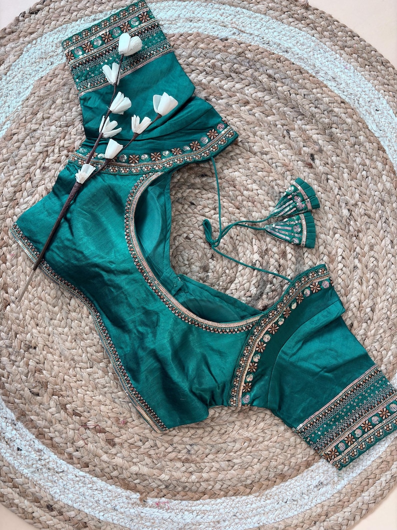 May include: Emerald green blouse with intricate embroidery along the neckline and sleeves. The blouse features a round neckline and short sleeves. The embroidery includes gold and copper-colored accents. The blouse is displayed on a woven jute rug.