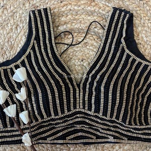 May include: Black sleeveless top with vertical rows of small, shiny, gold-colored beads. The top has a deep V-neck and a decorative beaded pattern at the bottom. A black string is visible at the back. The top is on a woven background.