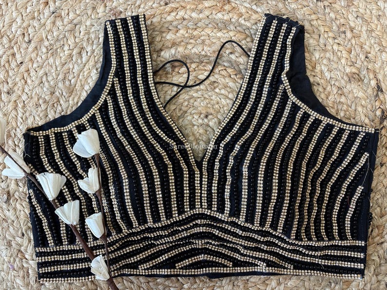 May include: Black sleeveless top with vertical rows of small, shiny, gold-colored beads. The top has a deep V-neck and a decorative beaded pattern at the bottom. A black string is visible at the back. The top is on a woven background.
