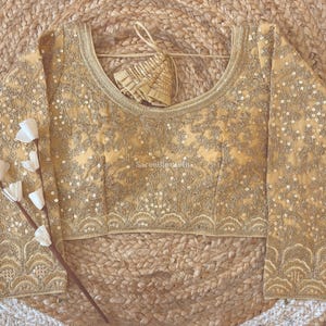 Könnte beinhalten: Eine goldfarbene Bluse mit langen Ärmeln, aufwendiger Stickerei und Paillettenbesatz. Die Bluse hat einen Rundhalsausschnitt und eine dekorative Bordüre am Saum und an den Manschetten. Ein passendes Accessoire hängt.