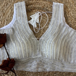 May include: White sleeveless top with a V-neckline, embellished with vertical rows of small, shiny beads. The top has a delicate, textured appearance and is paired with a decorative flower.
