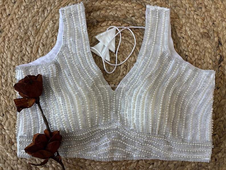 May include: White sleeveless top with a V-neckline, embellished with vertical rows of small, shiny beads. The top has a delicate, textured appearance and is paired with a decorative flower.