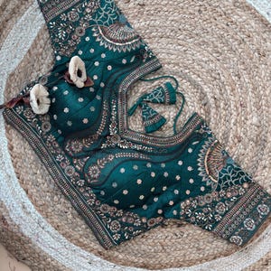 May include: An emerald green blouse with detailed embroidery and embellishments. The blouse features floral and paisley patterns in gold, pink, and white. The design includes a decorative neckline and sleeves, with matching tassels.