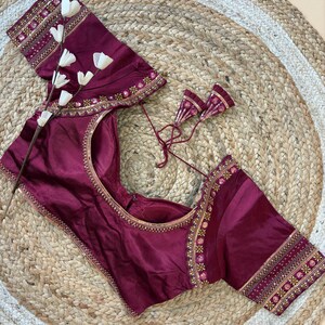 May include: A burgundy blouse with intricate gold embroidery along the neckline and sleeves. The blouse features a round neckline and short sleeves. The sleeves and neckline are adorned with detailed floral patterns. The blouse is displayed on a woven, circular mat.