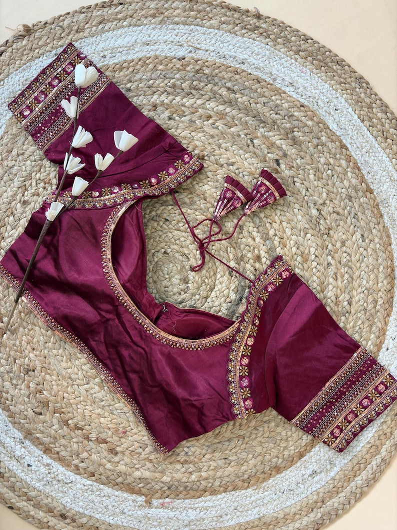 May include: A burgundy blouse with intricate gold embroidery along the neckline and sleeves. The blouse features a round neckline and short sleeves. The sleeves and neckline are adorned with detailed floral patterns. The blouse is displayed on a woven, circular mat.