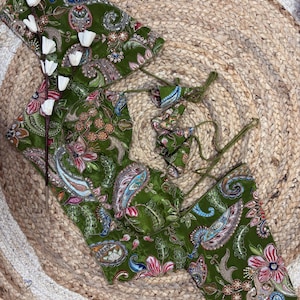 May include: A green paisley-patterned top and matching accessories. The fabric features a detailed design with pink, blue, and white floral and paisley elements. The items are displayed on a woven jute mat.