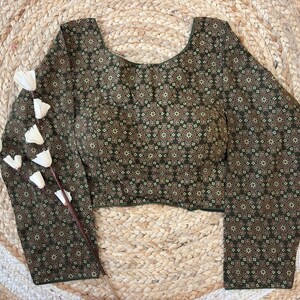 May include: A long-sleeved blouse with a dark green background and a repeating floral pattern in shades of brown and beige. The blouse has a scoop neckline and is displayed on a woven, circular mat with a sprig of white flowers.