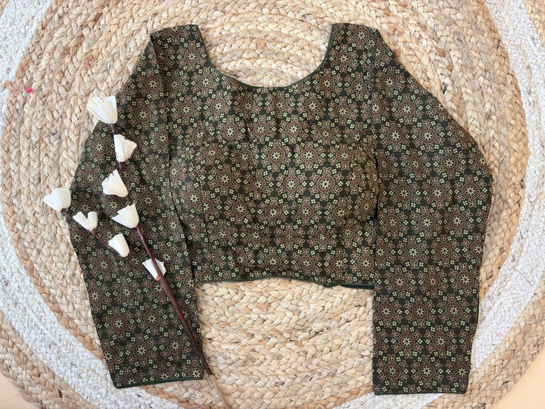 May include: A long-sleeved blouse with a dark green background and a repeating floral pattern in shades of brown and beige. The blouse has a scoop neckline and is displayed on a woven, circular mat with a sprig of white flowers.